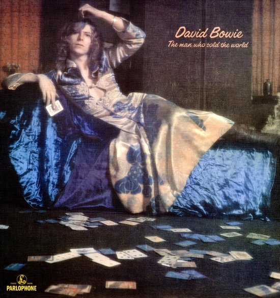 David Bowie op een bank in een bloemenjurk, met speelkaarten verspreid over de vloer. 'David Bowie The man who sold the world' in roze tekst.