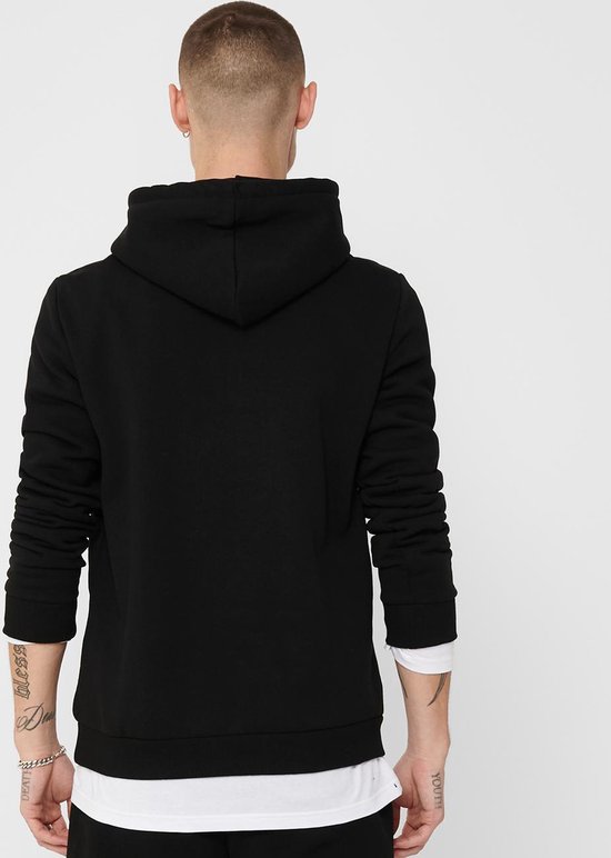 Man met geschoren haar en tatoeages op zijn armen draagt de achterkant van een zwarte hoodie.