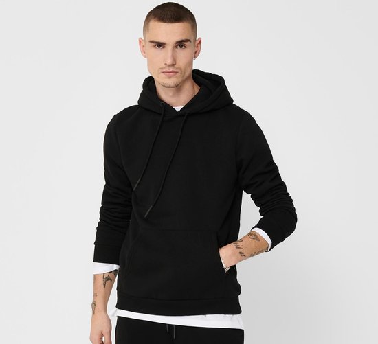 Man met kort geschoren haar en tatoeages, draagt een zwarte hoodie met capuchon en steekzak.