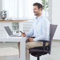 Man met een lichtblauwe blouse en beige broek zit aan een tafel, kijkend naar zijn telefoon.