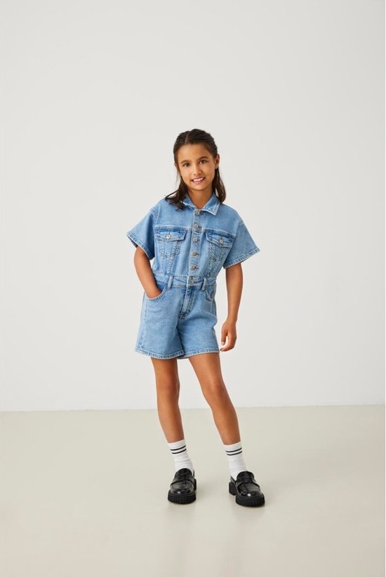 Meisje draagt een korte denim jumpsuit met korte mouwen, met een jeansblauwe wassing en omgerolde broekspijpen. Ze draagt witte sokken en zwarte schoenen.