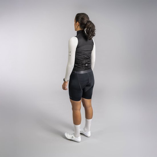 Vrouw met lange donkere haren in paardenstaart, draagt zwarte mouwloze fietsjas, lange witte mouwen, zwarte fietsbroek en witte sokken.