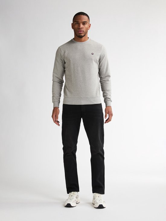 Man staat van voren, draagt een grijze sweater en zwarte jeans met witte sneakers.