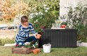 Zwarte opbergbox in een tuin, naast een vrouw die een plant verzorgt.
