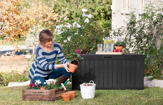 Zwarte opbergbox in een tuin, naast een vrouw die een plant verzorgt.
