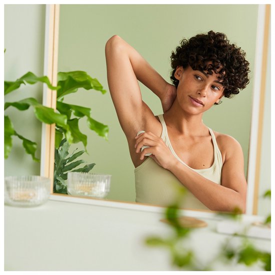 Een vrouw met krullend haar in een lichte badkamer past de deodorantstick toe onder haar arm.