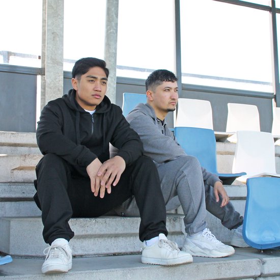 Twee mannen zittend op een trap bij een sportstadion. Ze dragen een Donnay trainingspak in het zwart en grijs.