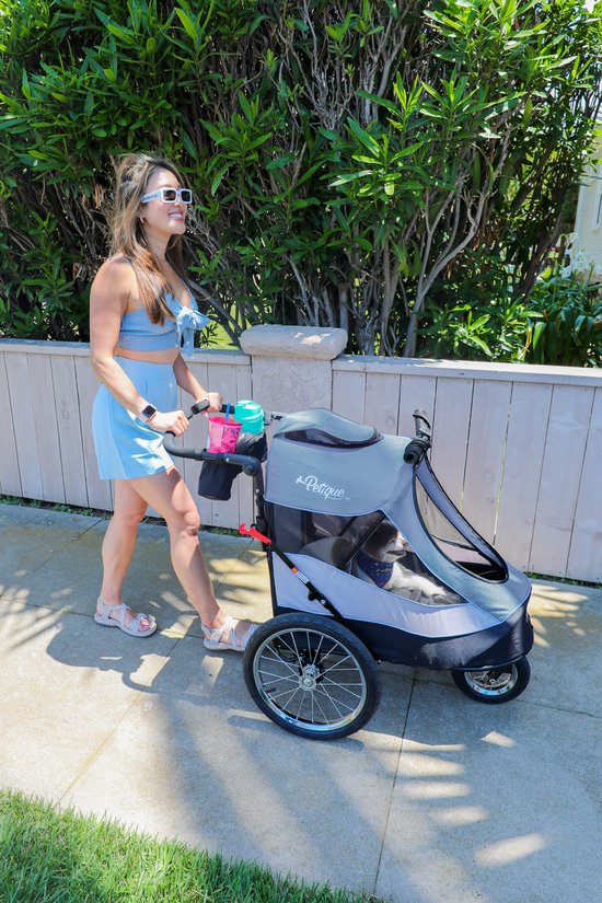 Vrouw in lichtblauwe outfit duwt een grijze hondenbuggy met een hond erin. De buggy heeft grote wielen en een beschermhoes.