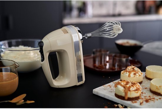 Crème handmixer van KitchenAid met garde op een donkere tafel, naast kleine taartjes.