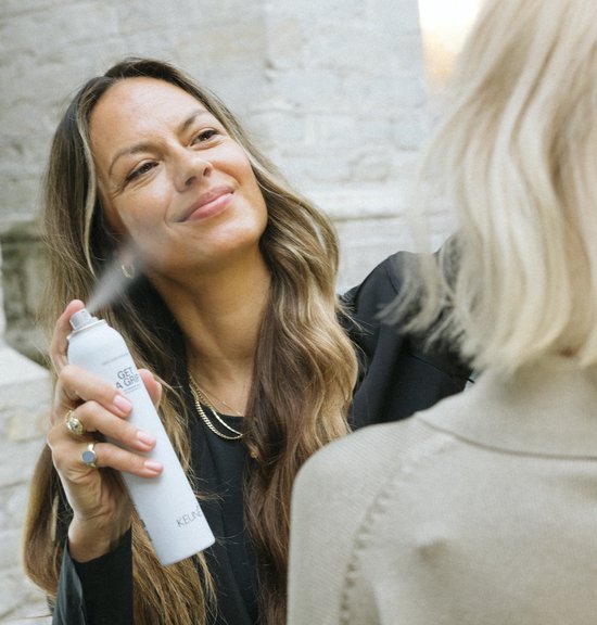 Een vrouw met lang bruin haar gebruikt Keune 'Get A Grip' spray op een ander model.