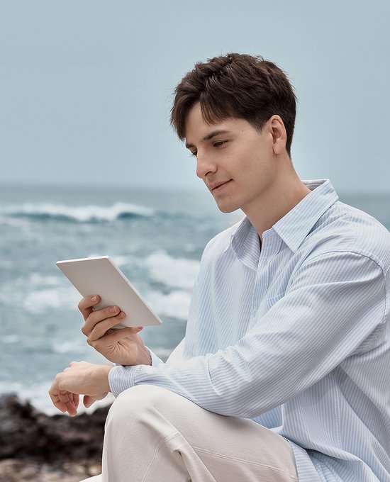Man met lichte blauwe gestreepte blouse kijkt naar een witte e-reader aan zee.