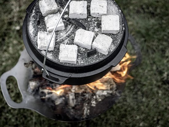 Zwarte Petromax Dutch oven met witte briketten op de deksel, boven een vuur met vlammen. Van bovenaf gezien, op gras.