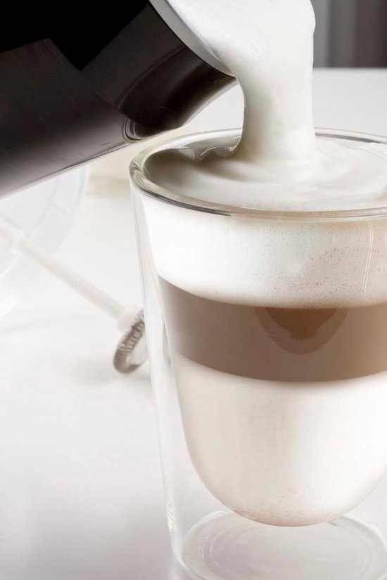 Zwarte melkopschuimer die dik melkschuim in een gelaagd glas latte macchiato giet.