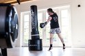Man traint met AJ-Sports bokszak van 175cm, draagt zwart shirt en korte broek met print.