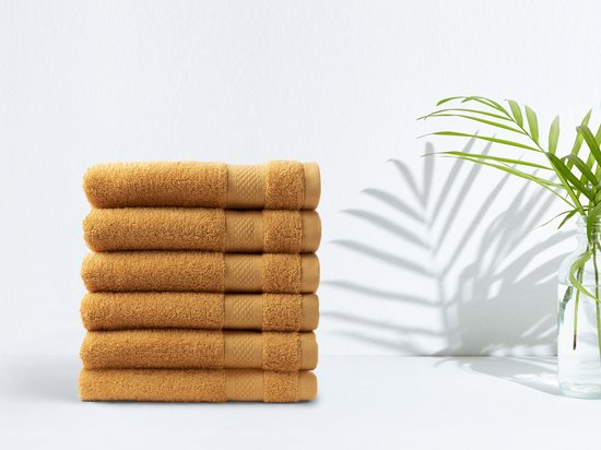 Stapel van zes okergele handdoeken op een witte achtergrond, met een groene plant op de achtergrond.