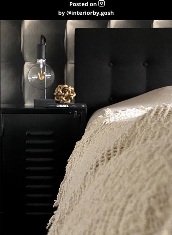 Zwarte tafellamp met grote gloeilamp op een nachtkastje naast een bed met beige sprei.