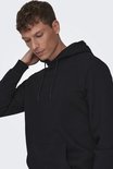 Man met bruine haren en een zwarte hoodie met capuchon, zachtjes zijn nek aanrakend.