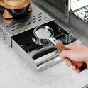 Een hand houdt een zilverkleurige portafilter met een bruin en zwart gestreept houten handvat boven een uitklaplade.