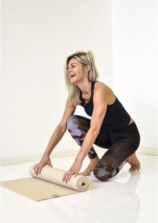 Een lachende vrouw met een patroon legging rolt een beige yogamat uit.