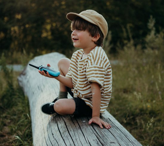 Jongetje met pet op een boomstam, met een blauwe Kidywolf KIDYTALK walkietalkie in de hand.