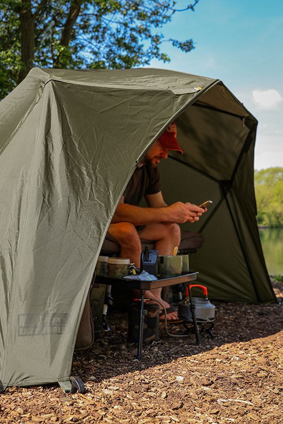 Man met pet, zittend in een olijfgroene visbrolly aan de waterkant, kijkend op zijn telefoon.