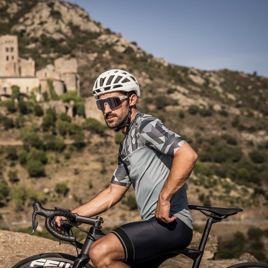 Mannelijke wielrenner met baard, draagt grijs met zwart fietsshirt en witte zonnebril, zittend op fiets.