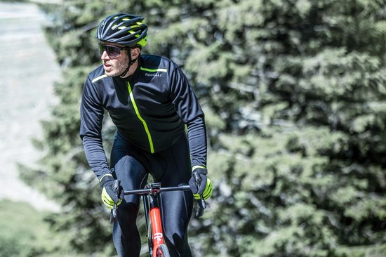 Man in zwart fietsjack met fluor gele rits en details, rijdend op een racefiets in bosrijke omgeving.