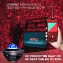 Een galaxy projector op een tafel, die rode nevels projecteert. Een smartphone met een muziek app is in beeld. De tekst 'De projector gaat op de beat van de muziek'.