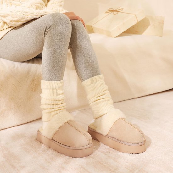 Model draagt beige suède pantoffels met dikke zool en crèmekleurige imitatiebontvoering bij grijze legging en sokken.