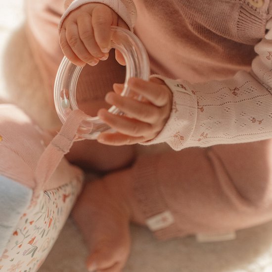Babyhandjes die een doorzichtige ring vasthouden, naast een zacht speelgoed. Baby draagt roze kleding.