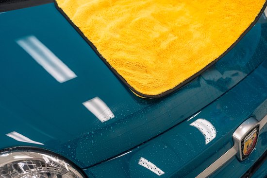 Een felgeleende microvezel droogdoek ligt op de teal blauwe motorkap van een auto, met waterdruppels.