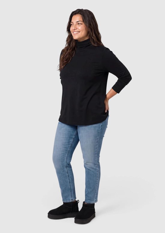 Zijaanzicht van een vrouw in een zwarte coltrui met lange mouwen, blauwe jeans en zwarte instappers.