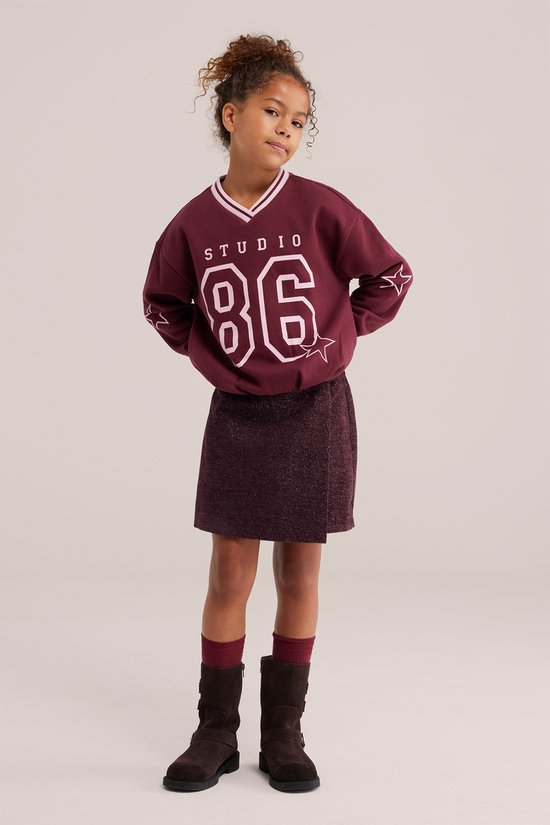 Meisje met krullend haar in een bordeauxrode sweater met 'STUDIO 86' en een bordeauxrode glitter wikkelrok.