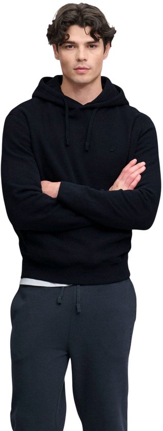 Man met gekruiste armen, draagt een zwarte hoodie met capuchon en een kleine geborduurde logo op de borst.
