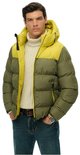 Model draagt een olijfgroene en gele gewatteerde Superdry jas met capuchon, open gedragen over een witte coltrui.
