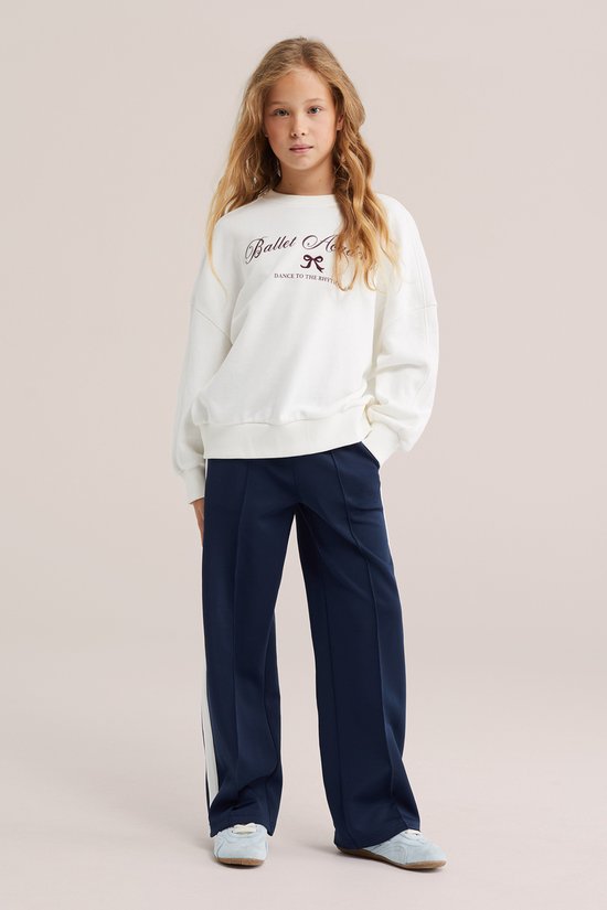 Model draagt een donkerblauwe wide leg broek met witte biezen en een witte trui met tekst "Ballet Academy".