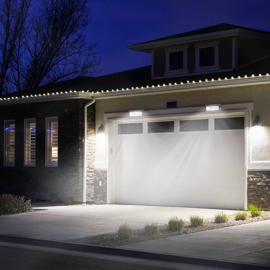 Een witte garagedeur wordt 's nachts verlicht door twee felle LED-lampen aan de gevel, met decoratieve lampjes langs de dakrand.