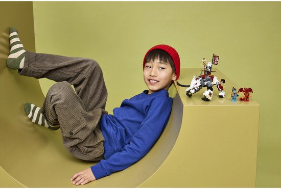 Een jongen met rode muts en gestreepte sokken zit naast een LEGO NINJAGO drakenplayset.