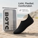 Zwarte waterschoen naast een doorzichtige verpakking met het BOTC-logo, op het strand. Beschrijving: Licht. Flexibel. Comfortabel. Perfect voor water & strand.