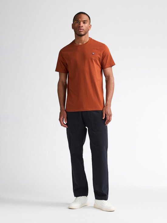 Man model in een effen oranje t-shirt met korte mouwen en navy broek. Witte sneakers.