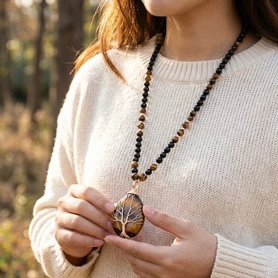 Close-up van een vrouw in wit gebreide trui met een lange ketting van zwarte en tijgeroogkleurige kralen, met een tijgeroog hanger in boomvorm.