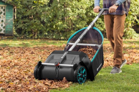 Een man duwt een Gardena bladverzamelaar met een zwarte opvangzak met turquoise rand over een gazon met herfstbladeren.
