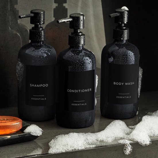 Drie natte, zwarte pompdispensers met schuim en zeep. Labels: SHAMPOO, CONDITIONER, BODY WASH.