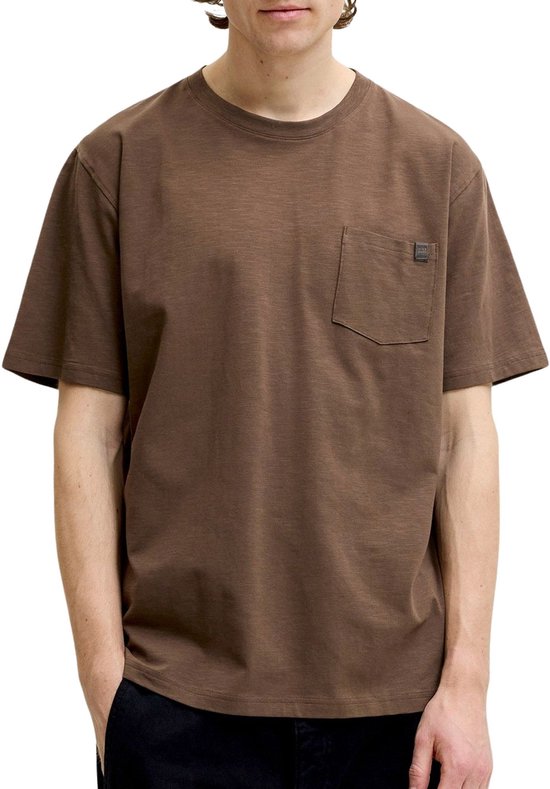 Man met handen in zakken draagt een bruin T-shirt met borstzakje.