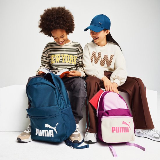 Twee kinderen, een jongen en een meisje, zitten naast elkaar met rugzakken. De jongen links heeft een donkerblauwe Puma rugzak. Het meisje rechts heeft een paarse en roze Puma rugzak.