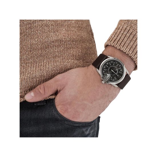 Man met bruine trui en spijkerbroek draagt een zilverkleurig Diesel horloge met donkerbruine leren band.
