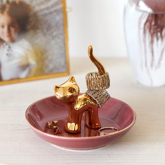 Gouden kattenbeeldje met ringen om de staart, in een roze schaaltje met gouden rand. Foto op de achtergrond.