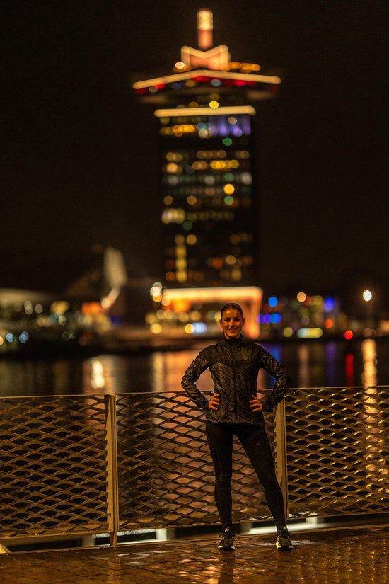Vrouw met handen in de zij, draagt een zwarte Rogelli hardloopjas met reflecterende details, 's nachts in de stad.