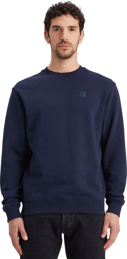 Man met donkerblauwe sweater met ronde hals en een vierkant logo op de borst. Een zwarte jeans is zichtbaar.