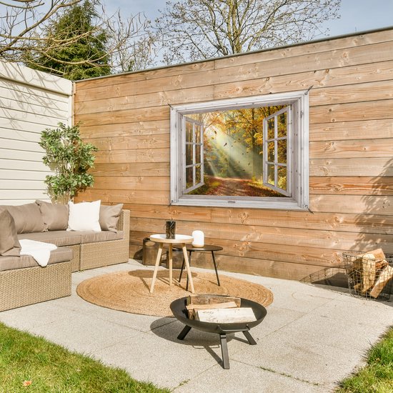 Een open houten raam met een bosweg in herfstkleuren, hangend aan een houten schutting naast een loungeset en vuurplaats.